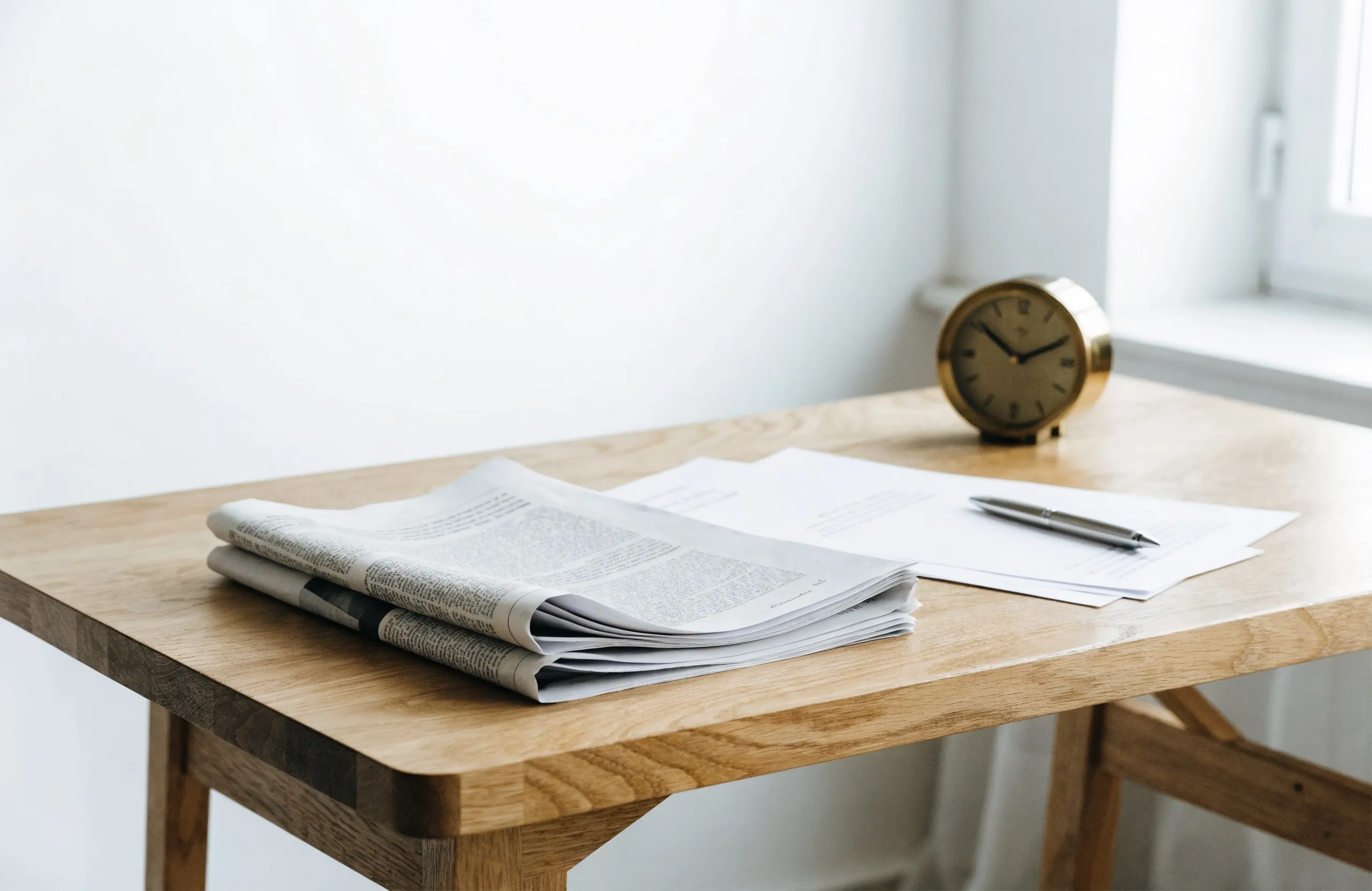 Folded newspapers, crisp letterhead pages, a silver pen and a brass desk clock on light oak in bright daylight